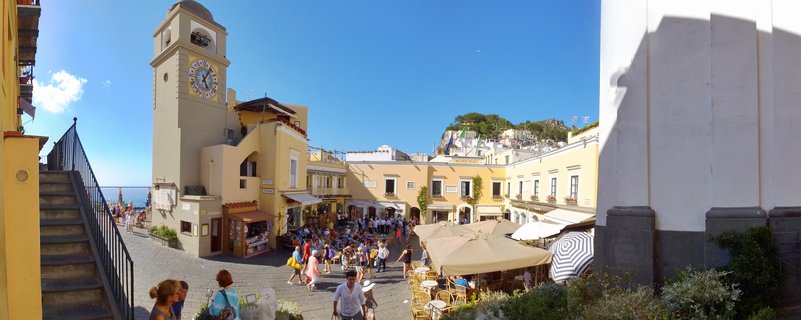 Aerial view of a crowded town square in Capri with a tall clock tower, yellow buildings, outdoor café umbrellas, and tourists walking on stone pavement under clear blue sky