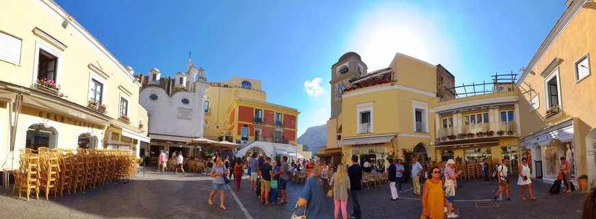 Wide panoramic view of Capri's main square with tourists walking among yellow and ochre buildings, outdoor cafes, clock tower, and blue sky with bright sun
