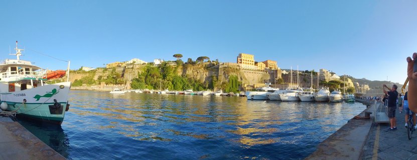 Panoramic view of Marina Grande harbor in Capri with white yachts docked in blue water, a ferry boat on the left, people on the stone quay, and yellow buildings on the cliffside under clear blue sky