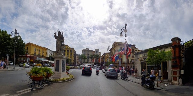 Street view of an Italian town square with a religious statue on a pedestal, colorful buildings, cars, scooters, and pedestrians under cloudy skies