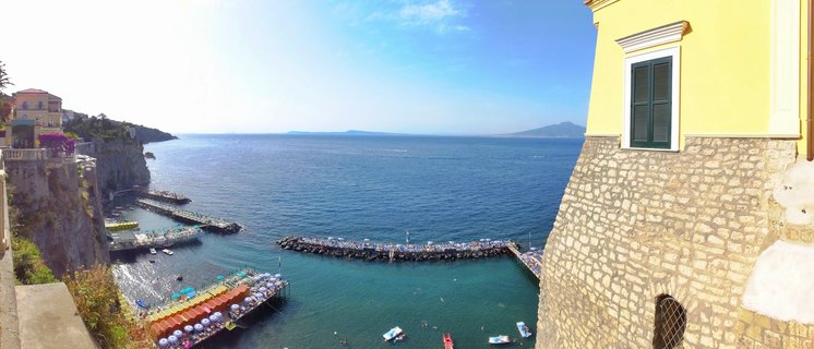 Panoramic coastal view of Sorrento Italy showing Mediterranean Sea with beach club platforms, colorful umbrellas, boats, yellow building with green shutters, and stone cliffs under clear blue sky