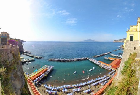 Aerial view of a crowded beach in Sorrento with rows of blue and white umbrellas, stone piers extending into turquoise water, yellow buildings on cliffs, and distant island on the horizon