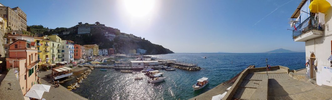 Panoramic view of Sorrento harbor showing colorful buildings along the waterfront, boats in blue Mediterranean water, and hills under bright sunny sky