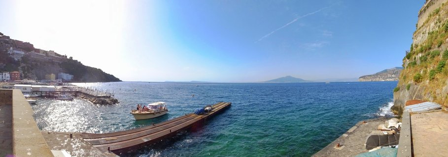 Panoramic view of Sorrento harbor in Italy showing a boat at wooden pier, turquoise Mediterranean water, cliffside buildings, and distant island under clear blue sky