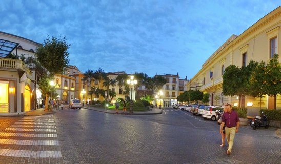 A panoramic view of an Italian town square at twilight with cobblestone streets, illuminated street lamps, historic buildings, palm trees, parked cars, and pedestrians walking