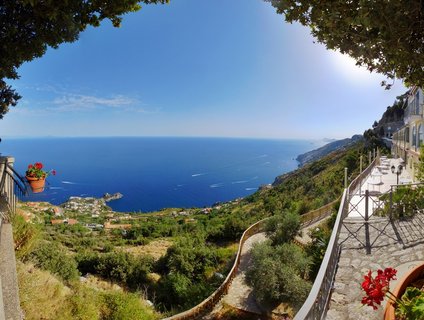 Wide coastal landscape showing a stone terrace with red flowers overlooking the blue Tyrrhenian Sea from San Lazzaro on the Amalfi Coast, with green hillsides, coastal villages, and boat wakes visible on the water under a clear sky.
