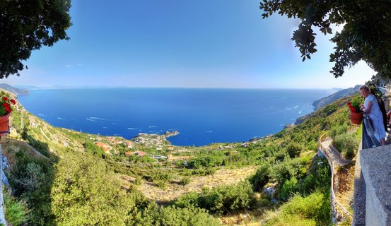 Panoramic view of Mediterranean coastline from elevated terrace showing deep blue sea with boat wakes, green hillside with red-roofed buildings, woman in light dress at railing, terracotta flower pots, and tree branches framing clear blue sky