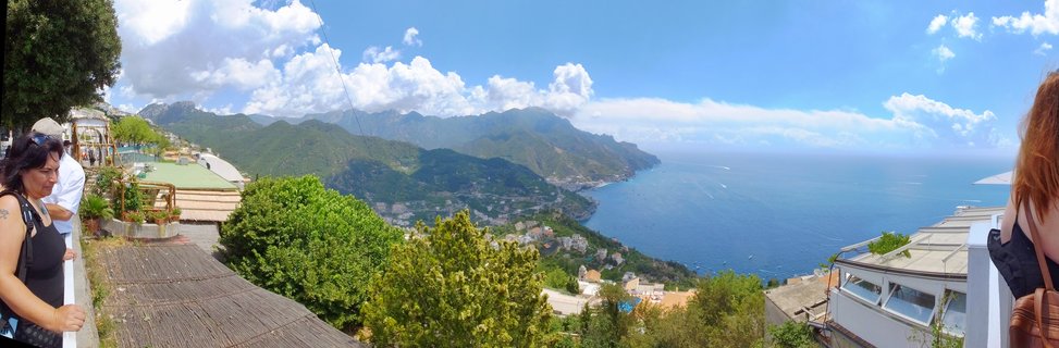 Panoramic landscape showing the Amalfi Coast from Ravello with mountains, Mediterranean Sea, coastal villages, and tourists at a viewpoint terrace