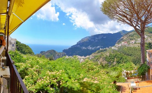 Scenic coastal view from a terrace in Ravello showing green hillsides, white buildings, mountains, and blue Mediterranean Sea under cloudy sky
