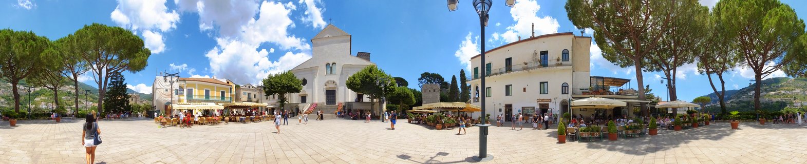 Wide panoramic view of a sunlit Italian town square with a white cathedral, outdoor restaurant seating with umbrellas, stone pavement, pine trees, and tourists walking under a blue sky with clouds
