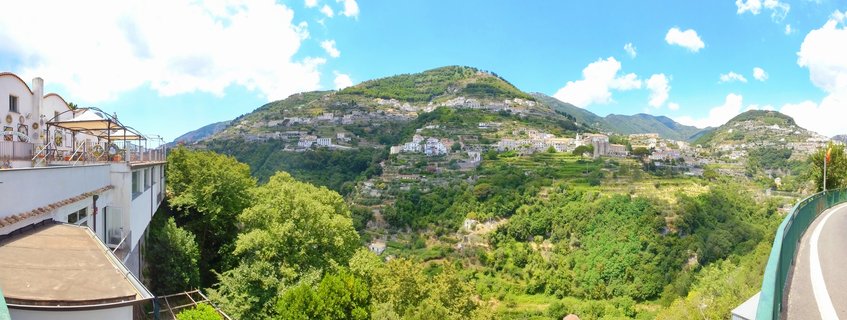 Panoramic view of white buildings scattered across green terraced hillsides in Ravello, Italy, with blue sky and clouds above