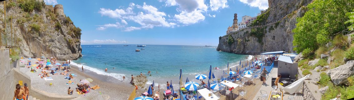 Aerial panoramic view of Marina Grande beach in Amalfi showing blue umbrellas, tourists on pebble beach, turquoise Mediterranean water, and buildings on limestone cliffs