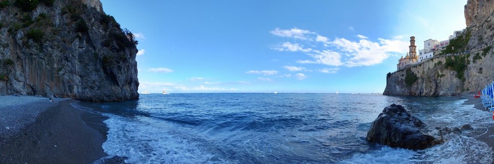 Panoramic coastal view showing a pebble beach between limestone cliffs, with a church bell tower and buildings on the right cliff face, blue sea with sailboats, and clear sky