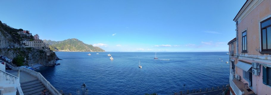 Panoramic coastal view showing the Amalfi coastline with buildings on cliffs, sailboats on blue Mediterranean water, and mountains in the distance under a clear sky