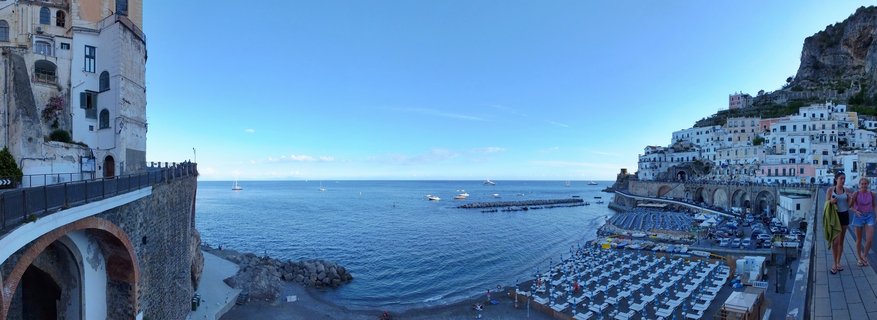 Panoramic coastal view showing a Mediterranean beach with organized umbrellas, boats in blue water, and white buildings built into cliffside in Atrani, Italy