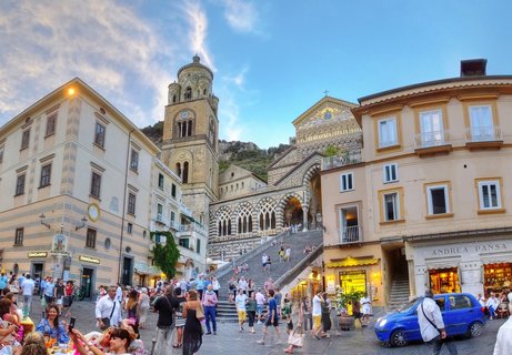 Wide view of Amalfi Cathedral with ornate striped facade and bell tower, grand staircase, tourists in piazza, surrounding buildings with shops, blue sky with clouds at dusk
