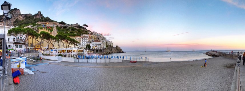 Panoramic sunset view of Amalfi town on Italy's coast with beach, blue umbrellas, hillside buildings, and sailboats on calm Mediterranean waters under pink and blue sky