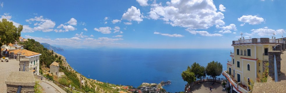 Panoramic view of San Lazzaro village on Italian coastline with yellow buildings, terracotta roofs, blue Mediterranean Sea, and scattered white clouds in summer sky
