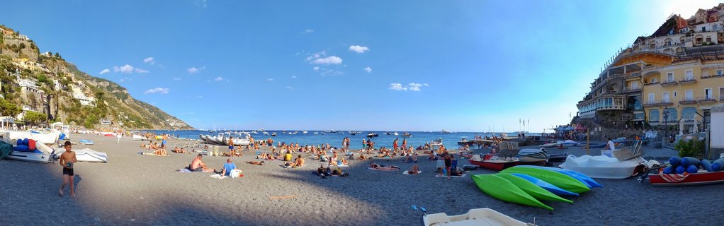 Wide beach scene showing tourists on dark pebbles at Positano's main beach with green and blue kayaks, boats on shore and in the water, colorful buildings on cliffs, and blue sky with clouds.