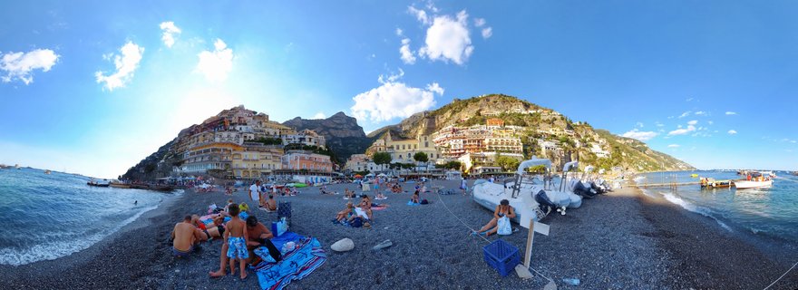 Panoramic beach view of Positano showing people on a pebble beach with colorful buildings built into a hillside, blue sky with clouds, and boats on the shore