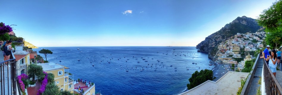Panoramic coastal view of Positano showing colorful buildings on steep cliffs overlooking the Mediterranean Sea with boats in the harbor and tourists on viewing terraces with purple flowers