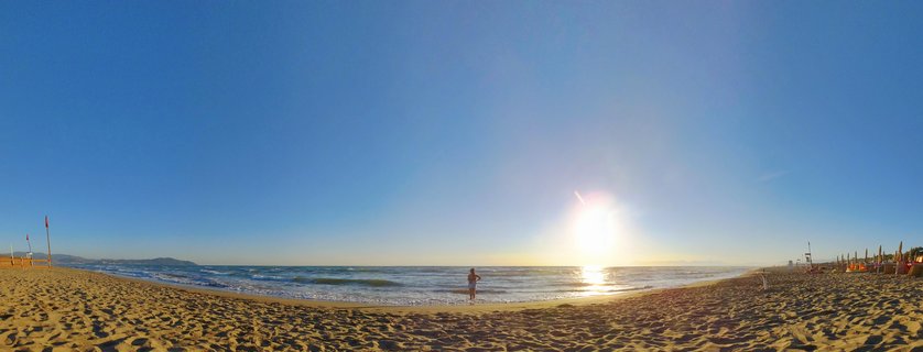 Wide panoramic beach scene showing a person standing at the water's edge during sunset, with golden sand in foreground, ocean waves, bright sun low on horizon, and clear blue sky over the Tyrrhenian Sea in Paestum, Italy