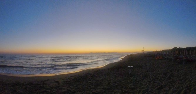 Panoramic beach scene at twilight showing the Tyrrhenian Sea with waves on sand, golden horizon, blue sky gradient, and beach resort structures on the right side near Paestum, Italy
