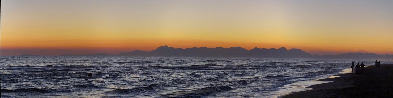 Panoramic seascape at sunset showing orange and yellow sky over dark blue water with waves, distant mountain silhouette on horizon, and people standing on beach at right side