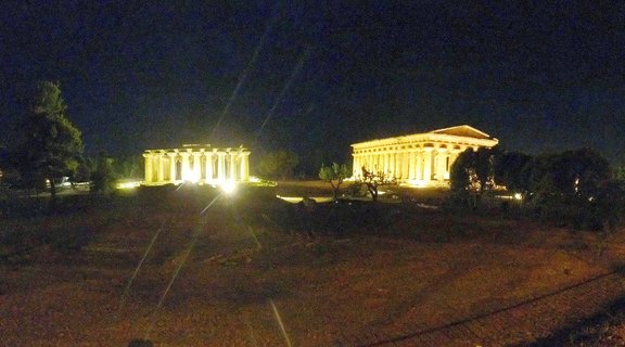 Two ancient Greek temples with Doric columns illuminated by warm artificial lighting against a dark night sky at the Paestum archaeological site in Italy