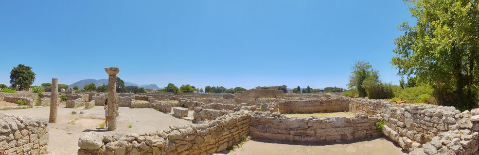 Panoramic view of ancient Greek archaeological ruins at Paestum, Italy, showing stone columns, wall foundations, and a visitor exploring the site under clear blue sky with distant mountains