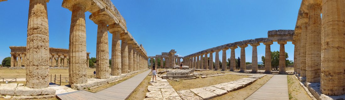 Ancient Greek temple ruins with rows of weathered stone columns under clear blue sky at Paestum archaeological site in Italy