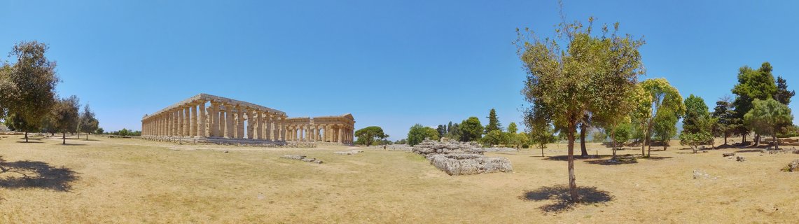 Ancient Greek temple with stone columns at Paestum archaeological site in Italy, surrounded by dry grass field and trees under clear blue sky