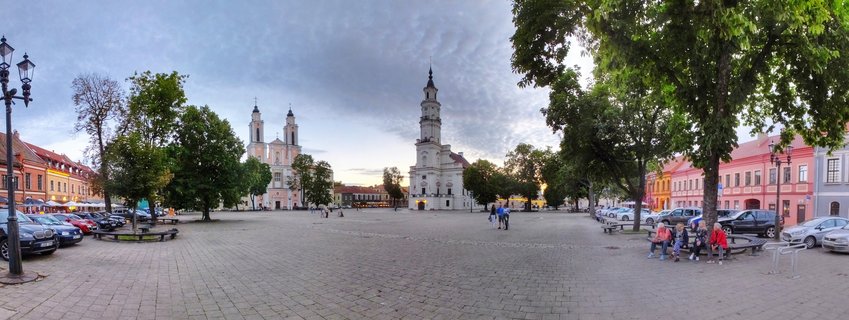 Wide panoramic view of Kaunas Town Hall Square showing cobblestone plaza, historic Church of St. Peter and St. Paul with twin towers, Town Hall tower, colorful buildings, trees, benches with people, and parked cars under cloudy evening sky