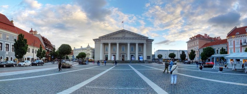 Wide panoramic view of Vilnius Cathedral Square showing the neoclassical cathedral with columns in the center, cobblestone plaza with white geometric patterns, pedestrians walking, and historic buildings with red roofs on both sides under a cloudy evening sky