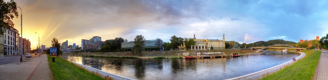 Wide panoramic photograph of Vilnius city at sunset showing the Neris River with buildings on both banks, Gediminas Castle Tower on a hill, bridge, walkways, trees, and dramatic sky with rainbow