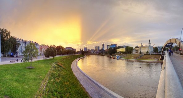 Panoramic cityscape of Vilnius showing the Neris River reflecting golden sunset light, with the arched White Bridge on the right, historic and modern buildings along both banks, and dramatic sun rays breaking through clouds