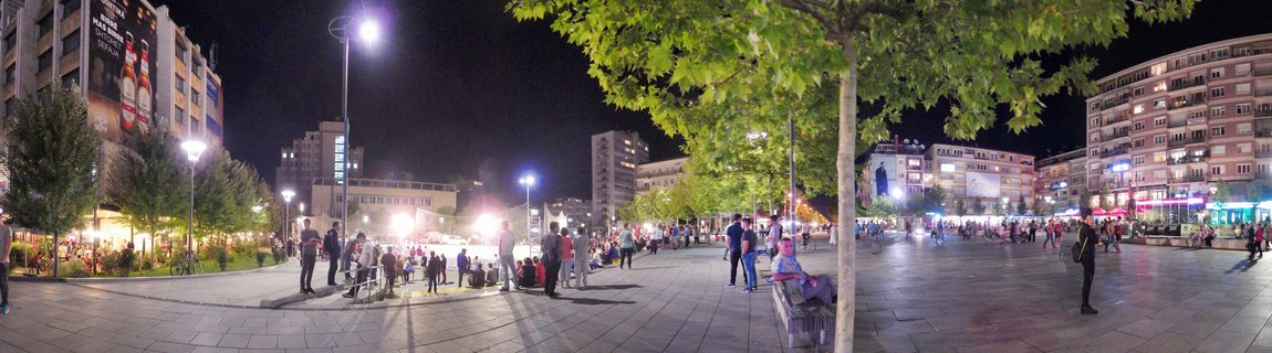 Night photograph of a busy urban square in Pristina with people walking and sitting on benches, surrounded by buildings and trees under street lighting