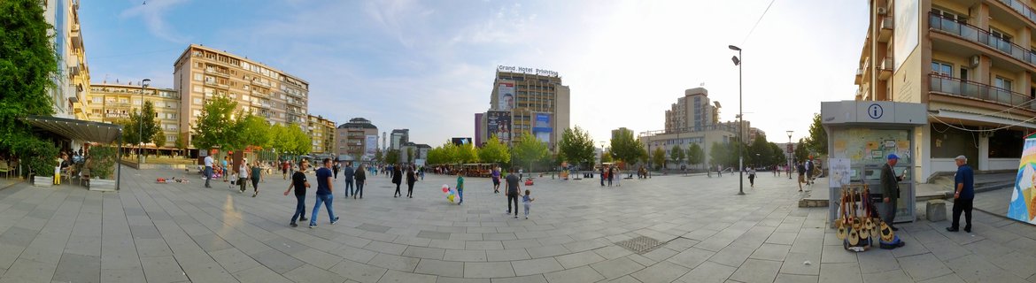Wide panoramic view of a city square in Pristina with people walking on paved ground, surrounded by multi-story buildings including Grand Hotel Prishtina, trees, and street infrastructure under a partly cloudy sky