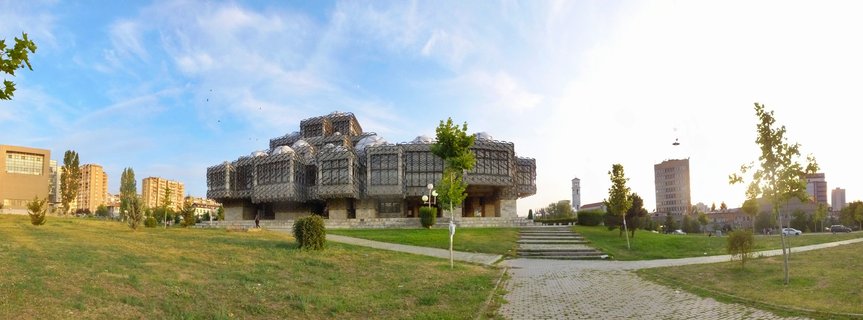 The National Library of Kosovo building with geometric concrete facade and multiple domes, surrounded by green lawn and trees under blue sky in Pristina