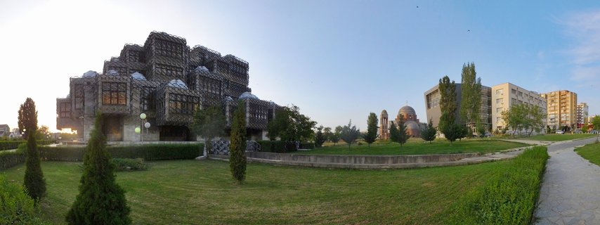 Wide panorama showing the National Library of Kosovo's distinctive brutalist concrete facade with geometric patterns and silver domes, green lawn in foreground, Orthodox cathedral dome visible in distance, clear blue sky