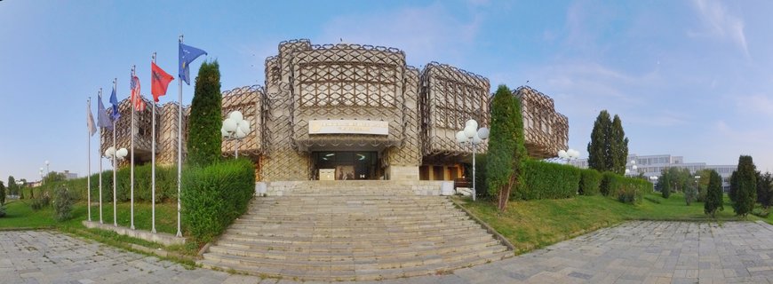 National Library of Kosovo building with geometric concrete facade, wide stone staircase, and multiple international flags on flagpoles against blue sky