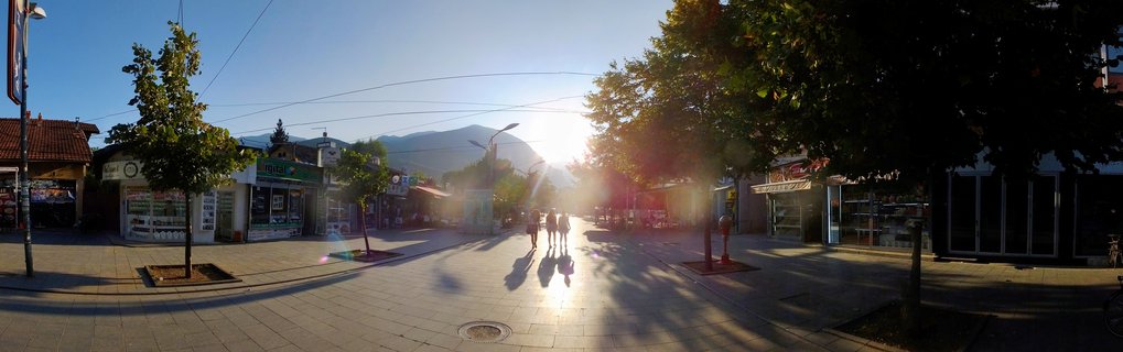 Three people walking on a paved pedestrian street in a town with shops on both sides, mountains in the background, bright sunset creating long shadows and lens flare