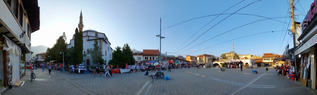 Wide panoramic view of a town square in Peja, Kosovo showing the Bajrakli Mosque with minaret, market stalls with colorful merchandise, pedestrians walking on cobblestone pavement, shops on both sides, and mountains in the distance under clear blue sky