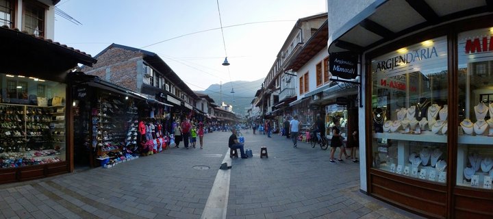 Wide street view of historic bazaar in Peja, Kosovo showing two-story traditional buildings with shops on both sides, pedestrians walking on paved street, jewelry and shoe store displays visible, mountains in background