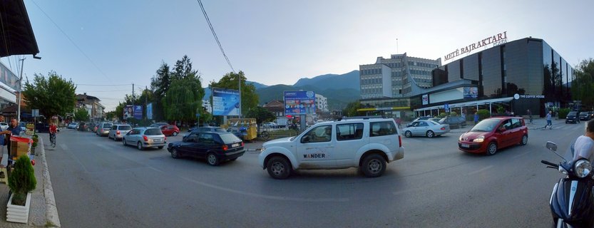 Wide panoramic street scene in Peja, Kosovo showing cars, a shopping plaza building with signage, cyclists, and mountains in the background under a clear sky