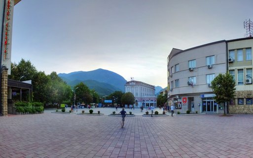 Wide panoramic view of a paved town square in Pejë, Kosovo with people walking and cycling, surrounded by buildings and trees with mountains in the background under an overcast evening sky