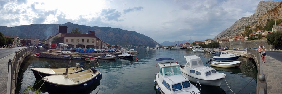 Panoramic view of Kotor harbor in Montenegro showing white and blue motorboats moored in calm bay water, with stone buildings, medieval architecture, and steep mountains in the background under cloudy sky