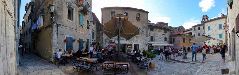 Wide view of stone plaza in Kotor Old Town with outdoor café seating, large umbrellas, tourists dining and walking, surrounded by historic limestone buildings with blue shutters and terracotta roofs under blue sky