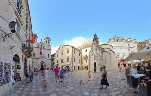Historic stone plaza in Kotor Old Town with tourists walking on checkered pavement, surrounded by medieval churches with bell towers, stone buildings with green shutters, outdoor café seating, and mountains visible in the background under blue sky