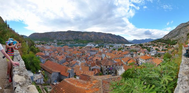 Panoramic photograph showing Kotor Old Town with red-tiled rooftops, stone buildings, surrounding mountains, and the Bay of Kotor with cruise ships visible in the distance under a partly cloudy sky
